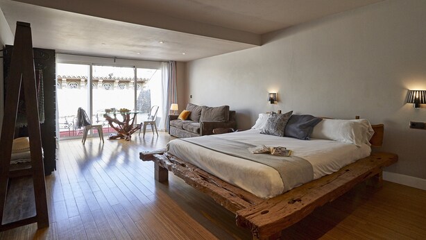 Een slaapkamer met een groot bed, een houten bankje, een sofa en een eettafel met stoelen.