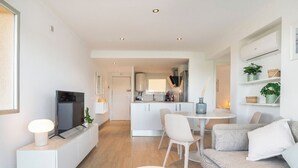 Ein modernes Wohnzimmer mit einer Couch, einem Couchtisch und einem Fernseher auf einem Lowboard. Im selben Raum befindet sich eine Küchenzeile mit Esstisch und Stühlen.