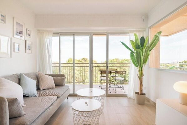A modern living room with a sofa, two small round tables, a potted plant, and a balcony with outdoor furniture.