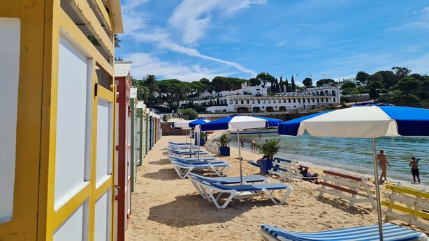 Ein Strand mit Liegestühlen und Sonnenschirmen, ein Gebäude mit weißer Fassade und einem strahlend blauen Himmel.
