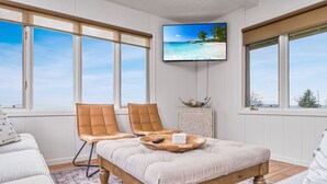 A room with a large bed, two leather chairs, a TV on the wall, and a view of the ocean.