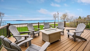 A deck with Adirondack chairs and a rectangular fire pit, overlooking a beach and grassy area.