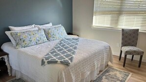 Un dormitorio con cama, almohadas, una silla y una ventana con persianas.