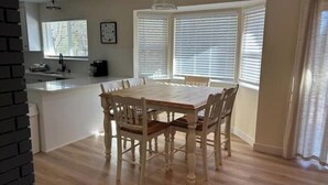 Un comedor con una mesa y sillas de madera, una cocina con pileta y mostrador, y ventanas amplias con persianas.