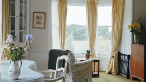 Una habitación con ventanal panorámico, una mesa con un jarrón de flores, una silla y vistas al exterior con vegetación.