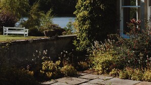 Un edificio de fachada de piedra, un jardín con un banco y vistas a un lago.