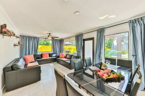 Un salon moderne avec un canapé d’angle, une table à manger en verre ornée de fruits et de grandes fenêtres avec des rideaux.