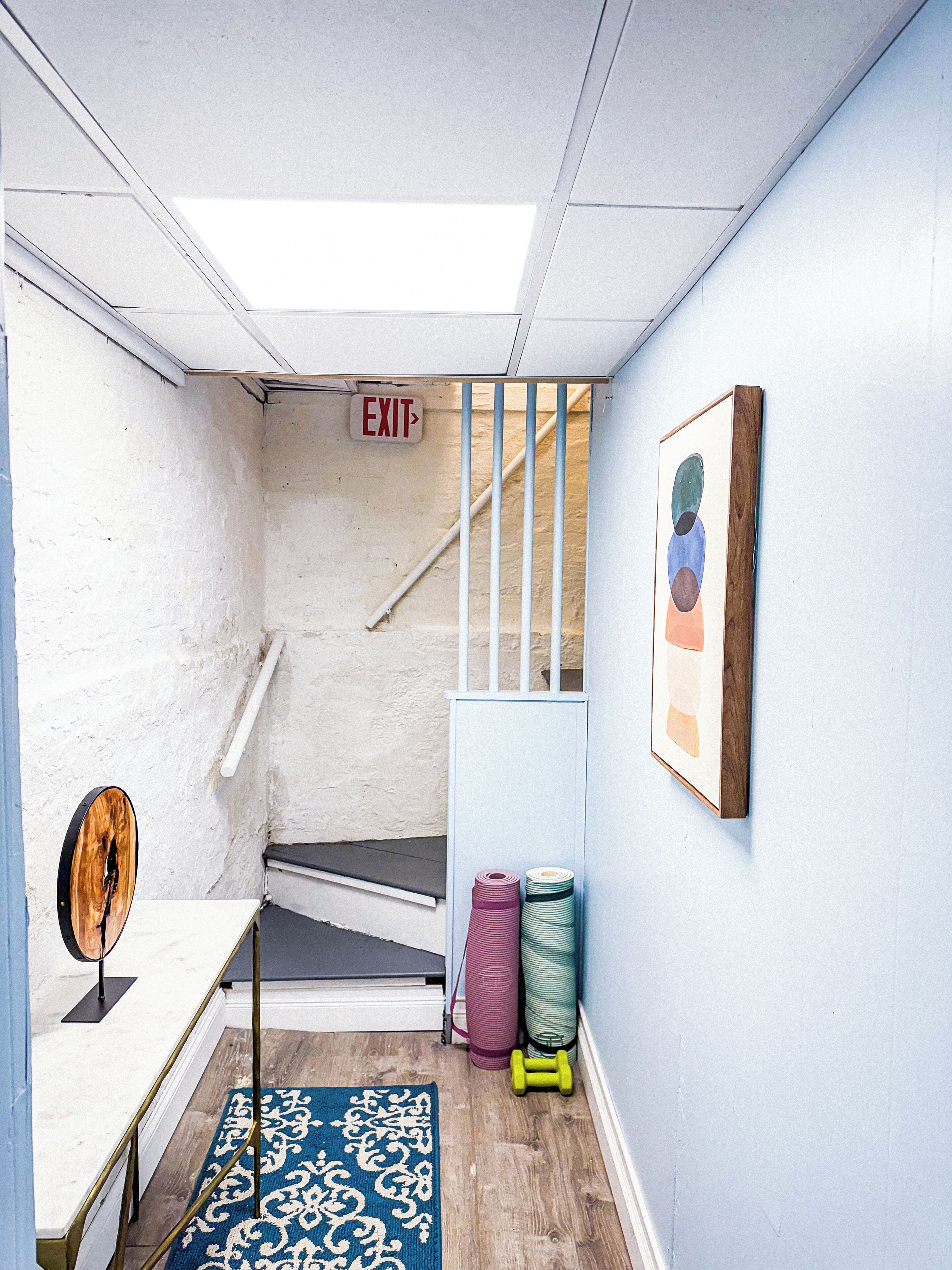 A compact room with a blue wall, a wooden floor, a blue patterned rug, a small table, a mirror, a framed picture, and a staircase with an exit sign.