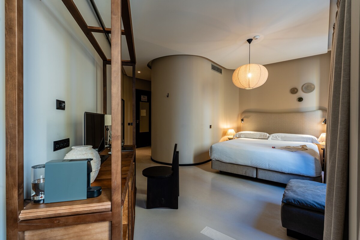 Habitación de hotel con cama, mesita de noche, lámpara y vista al pasillo.
