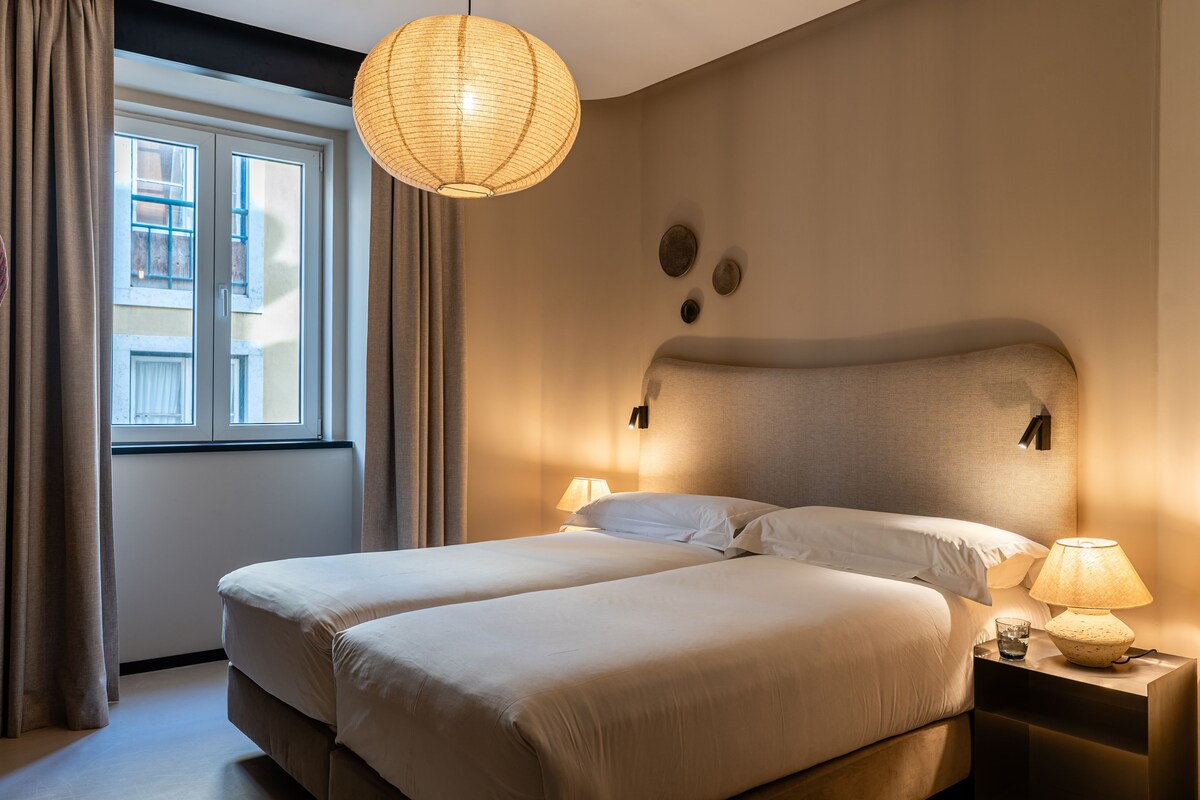 A double bed with two pillows, a bedside table with a lamp, and a window with curtains.