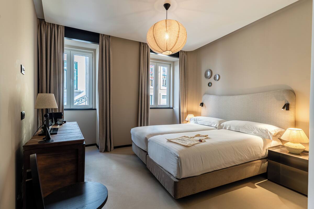 A hotel room with two beds, a desk with a lamp, a large window with curtains, and a hanging light fixture.