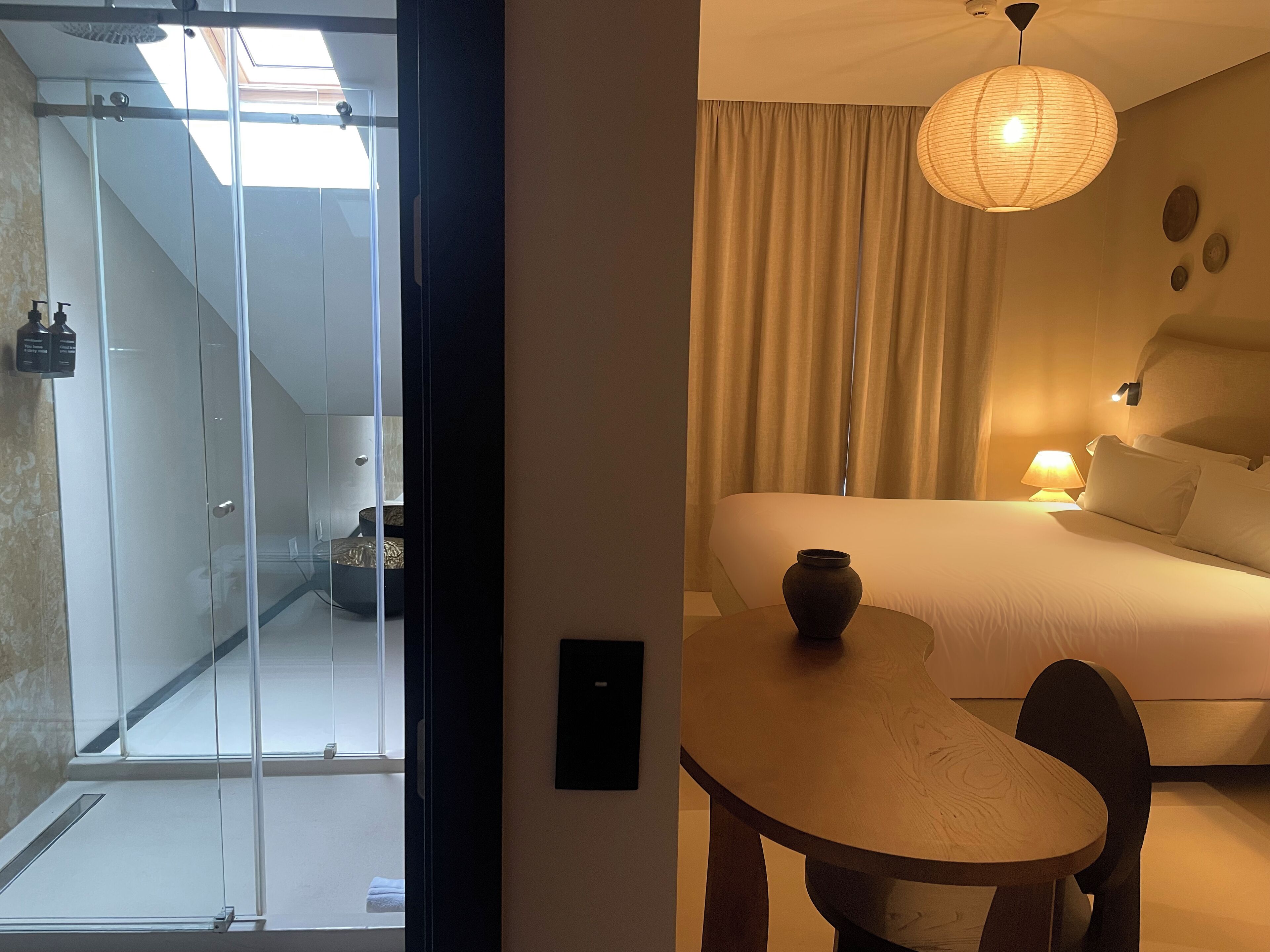 Quarto de hotel com cama, mesa de cabeceira, luminária e vista para um chuveiro em box de vidro.