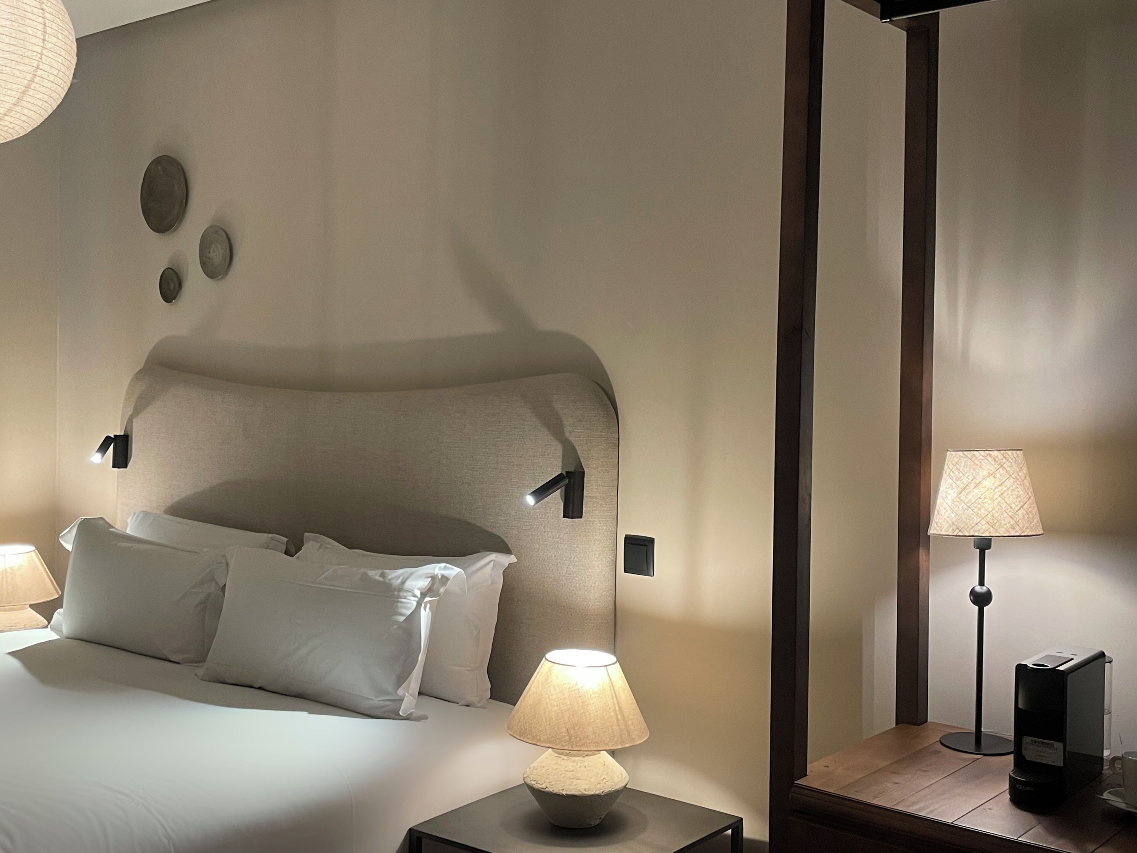 A neatly made bed with white linens, a bedside lamp, and a coffee maker on a wooden nightstand.