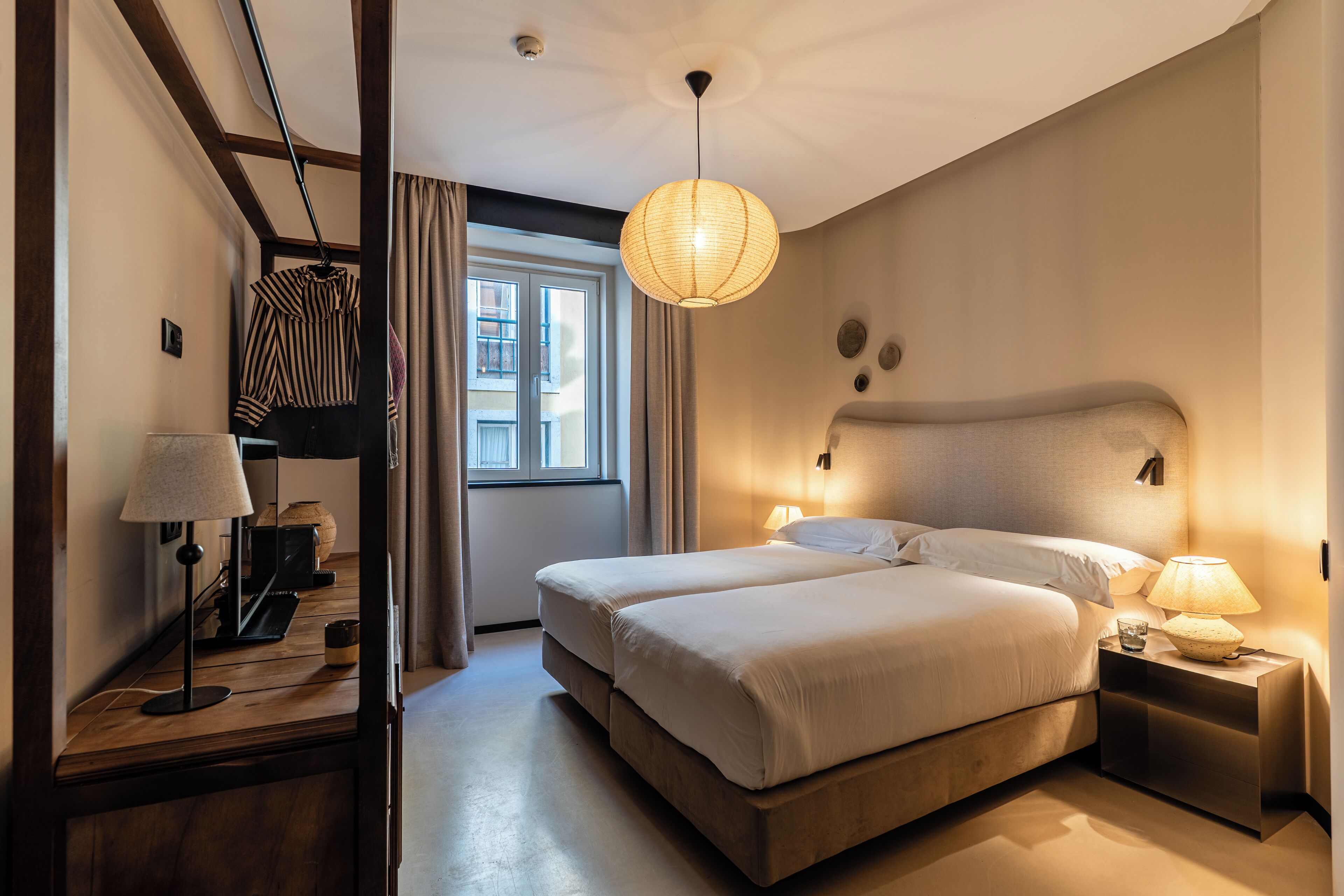 A hotel room with a large bed, bedside lamps, a hanging light fixture, a window with curtains, and a wooden floor.