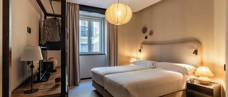 A hotel room with a large bed, bedside lamps, a hanging light fixture, a window with curtains, and a wooden floor.
