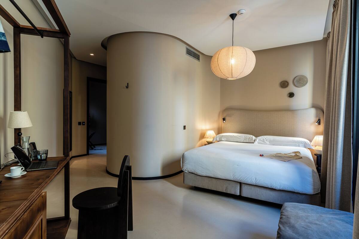 A modern hotel room with a curved wall, a bed with white linens, a desk with a laptop and a lamp, and a hanging light fixture.