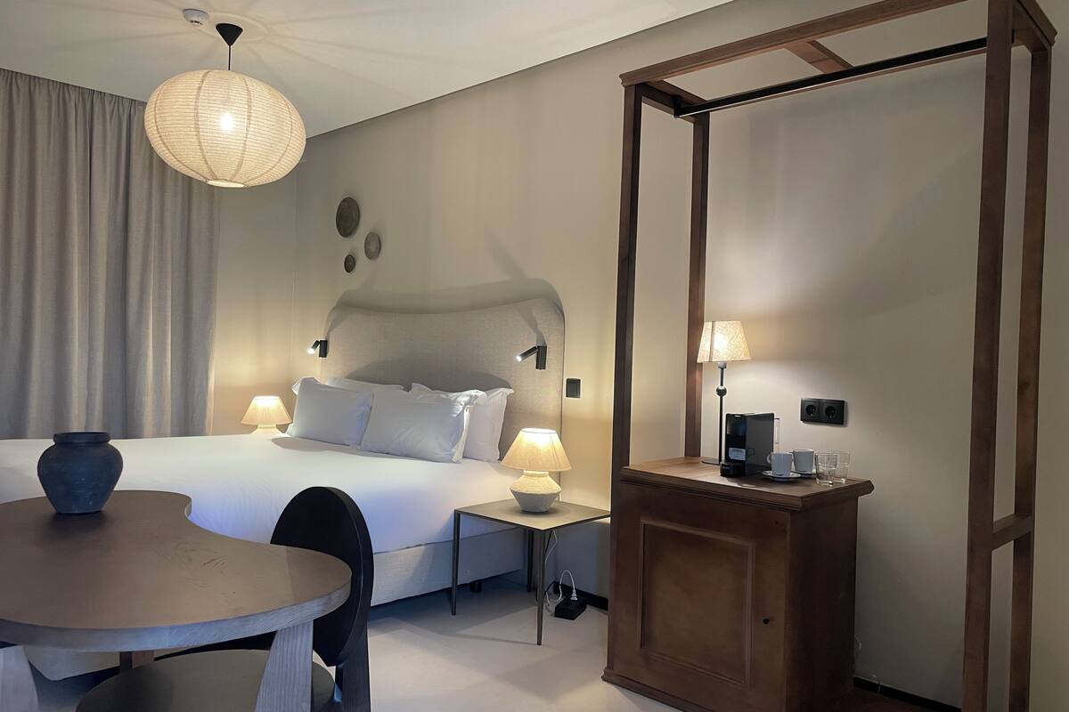 A hotel room with a bed, bedside tables, a small table, a chair, a lamp, a coffee maker, and a wooden cabinet.
