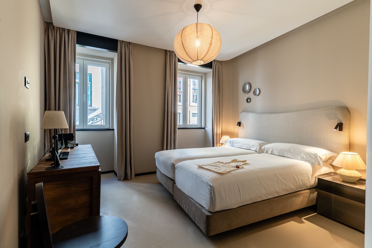 Habitación de hotel con dos camas, un escritorio, una silla, una lámpara y vistas al exterior.