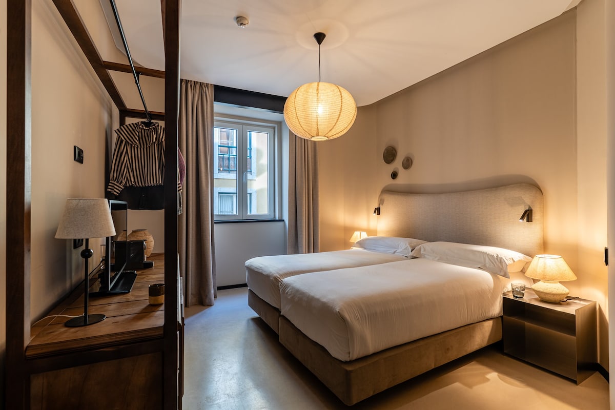 A hotel room with a bed, bedside lamps, a hanging lamp, a window with curtains, and a door.