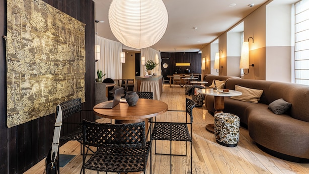 A modern hotel lobby with a round wooden table, black woven chairs, a large hanging light fixture, a brown sofa, and a large tapestry on the wall. - Casa Teva Lisboa by chic&basic (Lisbon)