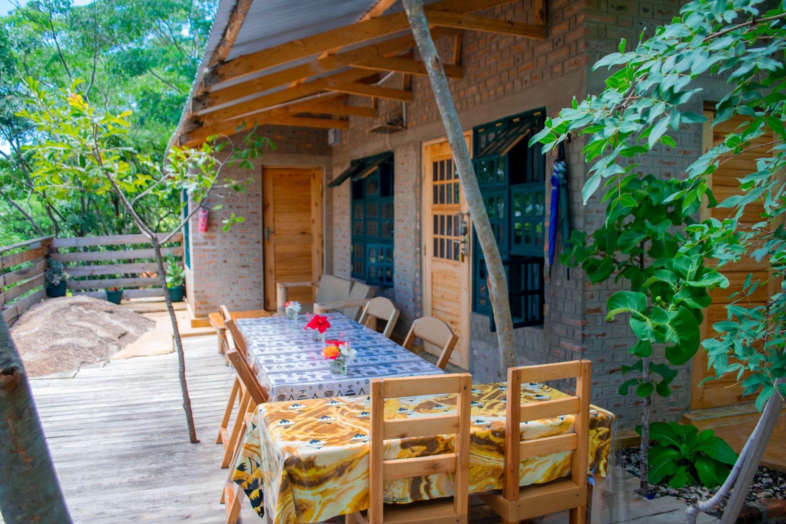 Une terrasse en bois avec une table et des chaises, entourée de verdure et bordée d’un bâtiment en briques.