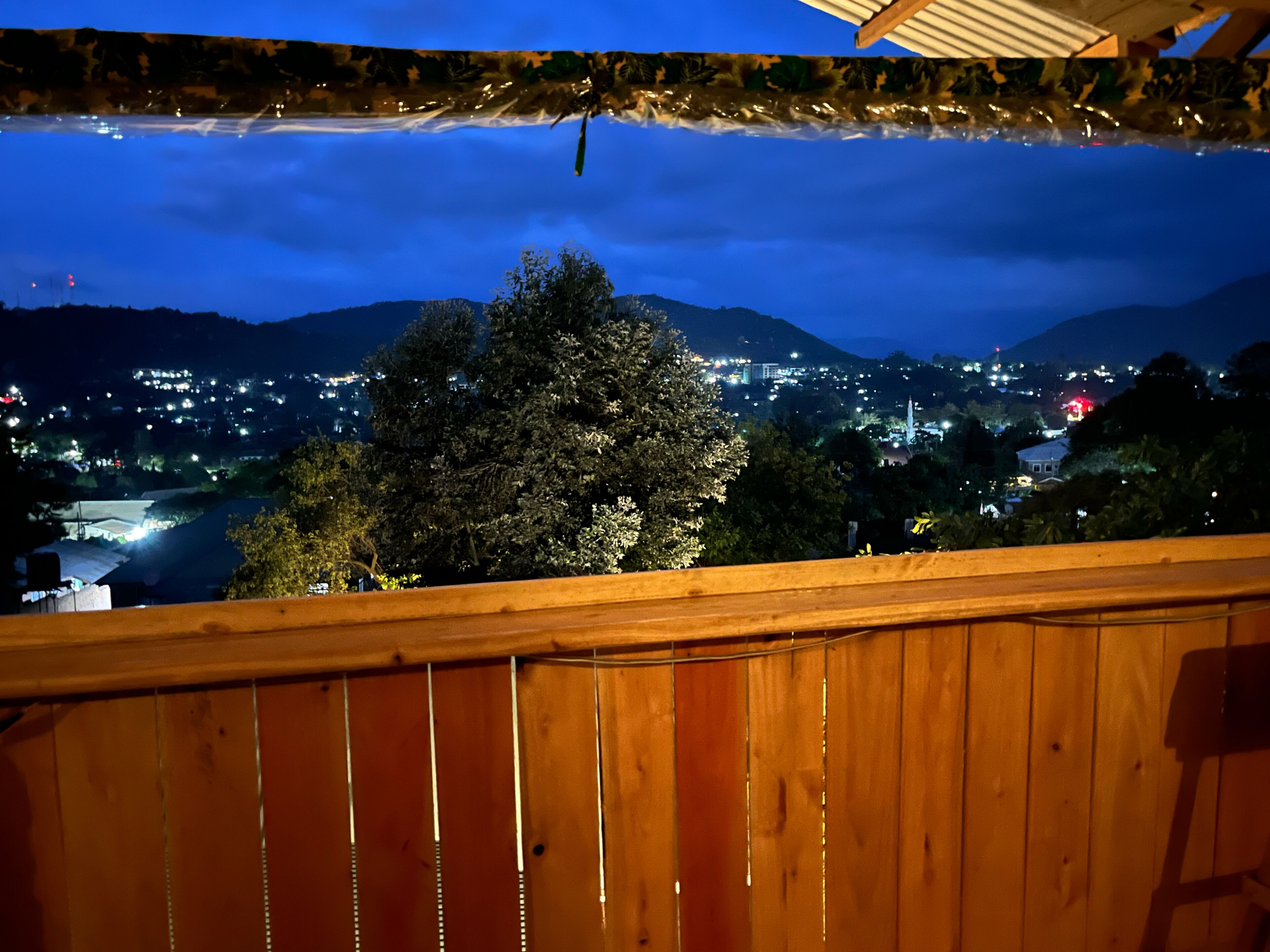 Un balcón de madera con vista a un paisaje urbano nocturno, con edificios iluminados y una montaña al fondo.