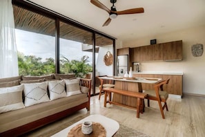 A modern living room with a sofa, dining table, and bench, a kitchen with wooden cabinets, and large windows with a view of greenery.