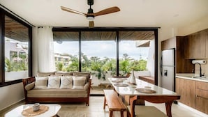 A modern living area with a sofa, dining table, and bench, large windows, and a ceiling fan. - Nasha Tropical Bliss Plunge Pool Beach Club Access (Tulum)