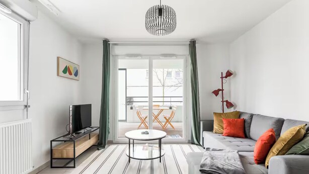 Ein modernes Wohnzimmer mit grauem Sofa, einem kleinen runden Tisch, einem Fernseher auf einem Schrank und Blick auf den Balkon.