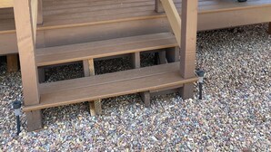 Eine Holzterrasse mit zwei Stühlen, einem Tisch, einem Grill und einem Fenster mit Jalousien.