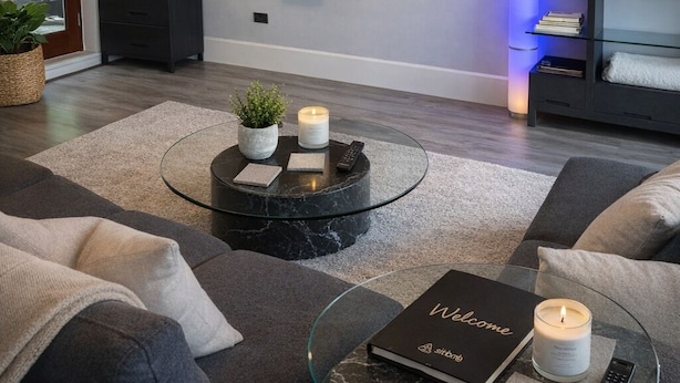Ein modernes Wohnzimmer mit einem Flachbildfernseher, einem runden Couchtisch mit Buch und Kerze, einer Couch und einer Vitrine mit Dekoration.