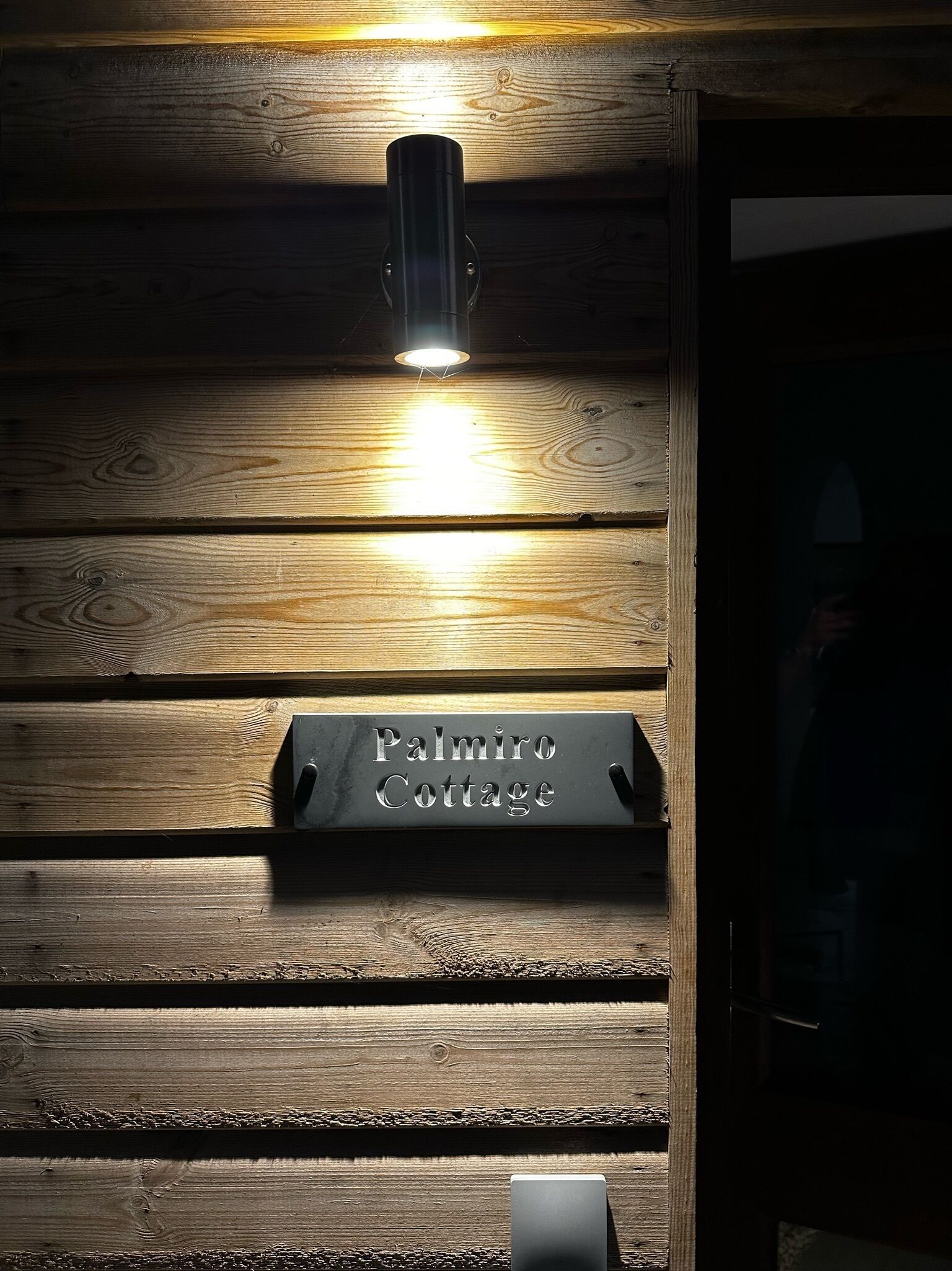Un mur en bois sur lequel est fixée une enseigne indiquant « Palmiro Cottage », et un luminaire placé au-dessus.