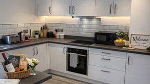 A modern kitchen with white cabinets, a built-in oven, a microwave, and a coffee maker. There is a bar stool and a trash can.