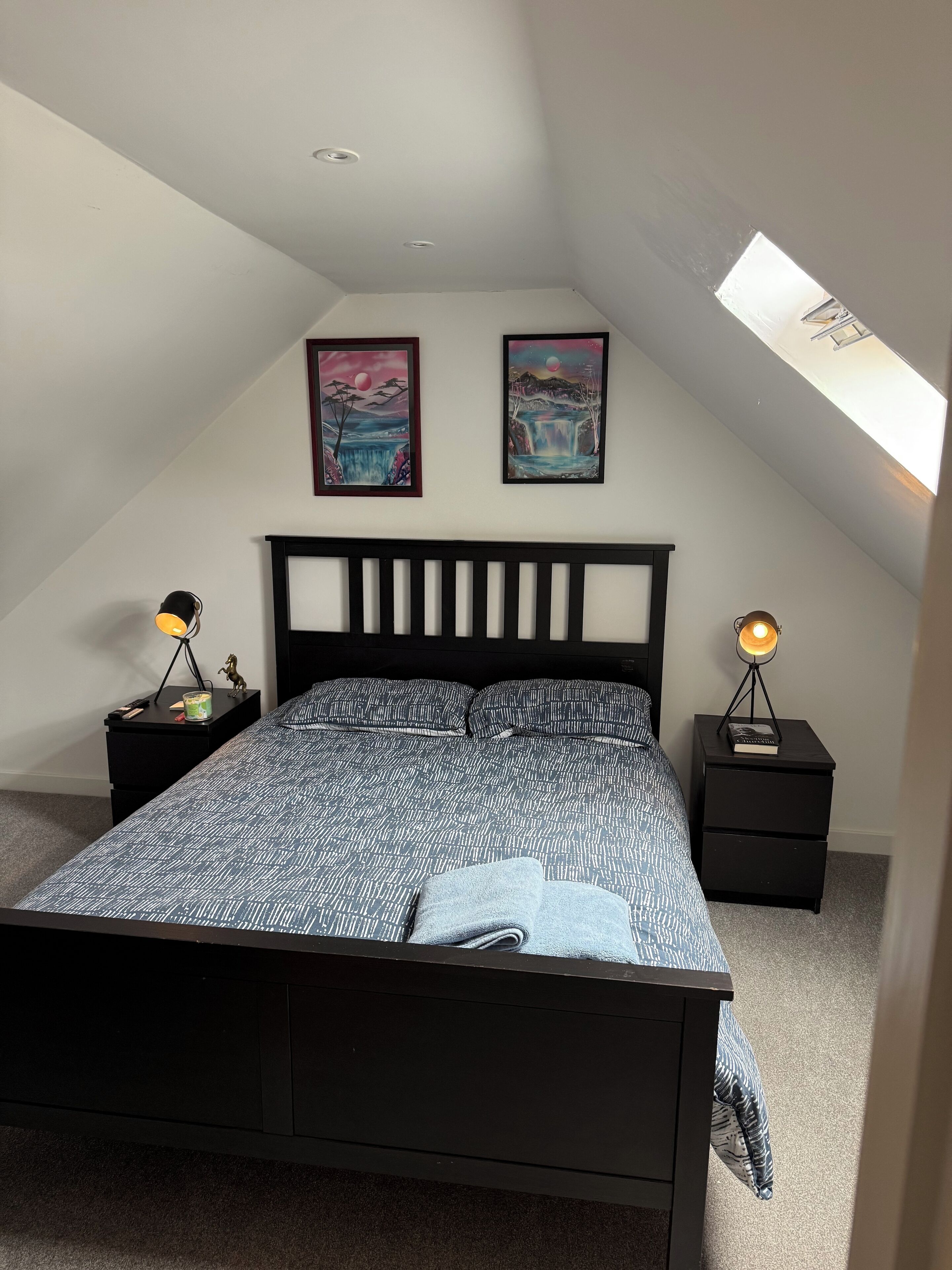 A bedroom with a sloped ceiling, a bed with a black headboard, two wall-mounted paintings, and two bedside tables with lamps.