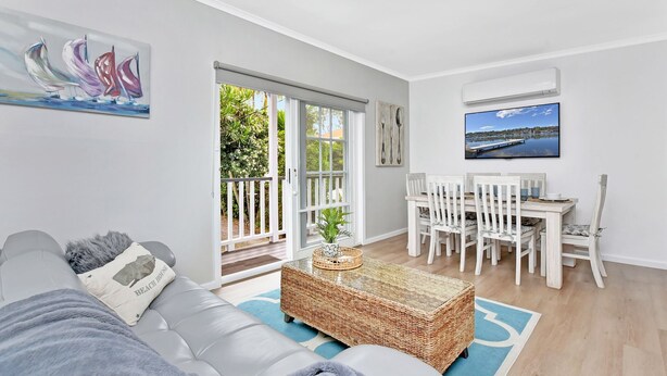 A modern living room with a sofa, a dining table with chairs, a TV, and a balcony with a view of greenery. - The Palms Retreat - Top Floor Apartment style (Nelson Bay)