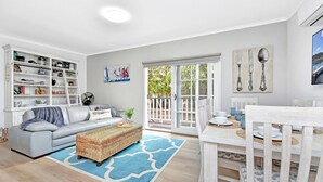 Uma sala de estar moderna com um sofá cinzento, uma mesa de jantar para quatro pessoas e uma grande janela com vista para o verde.
