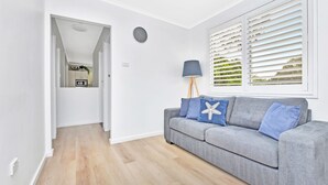 A modern living room with a grey sofa, a lamp, and a view through white blinds. - The Palms Retreat - Top Floor Apartment style (Nelson Bay)