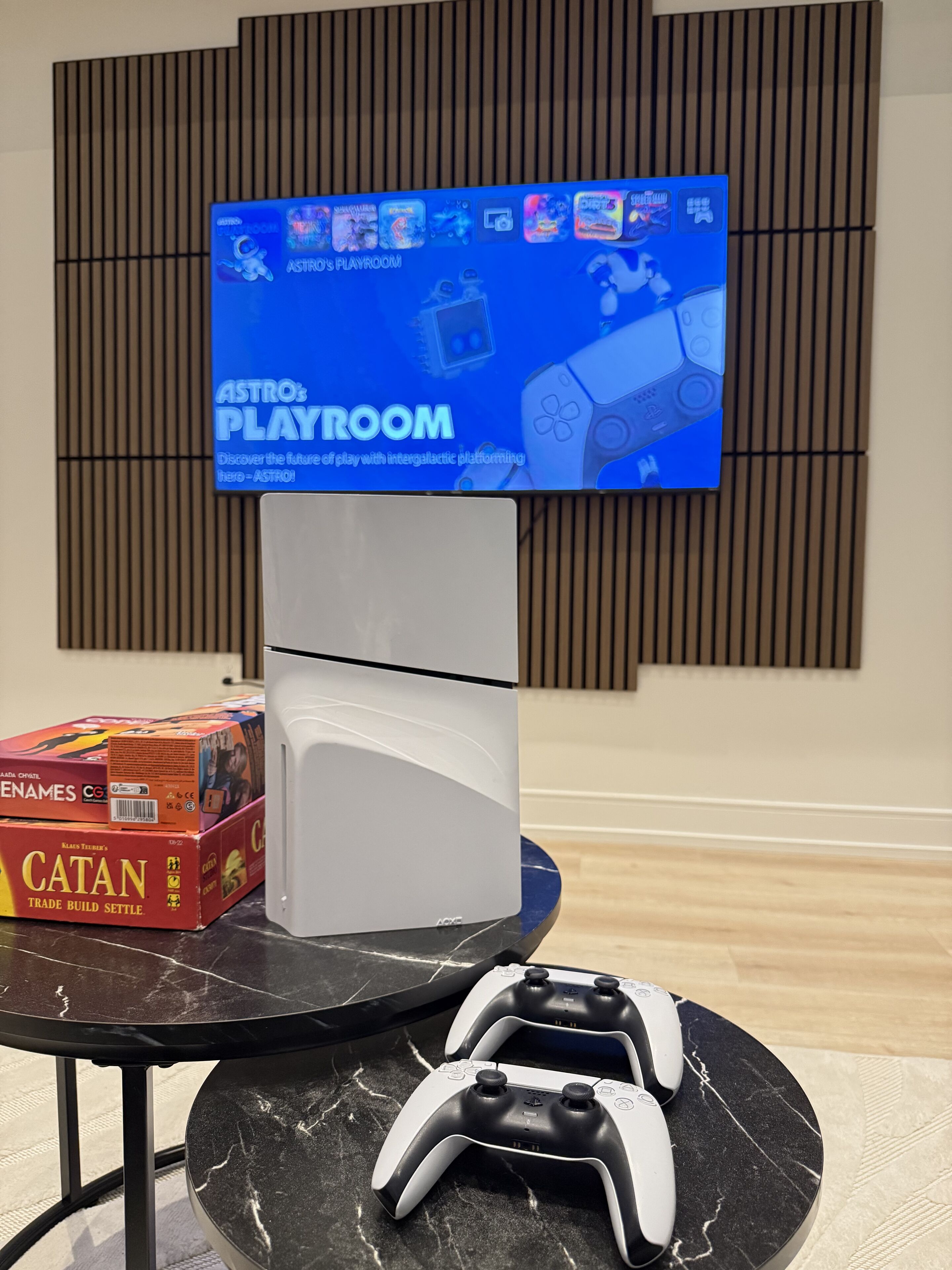 Un'area giochi con una TV che mostra 'ASTRO'S PLAYROOM', una console di gioco bianca e due controller su un tavolo di marmo.