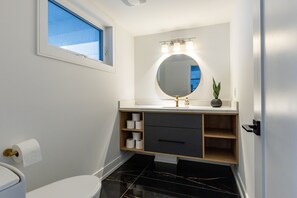 A modern bathroom with a white vanity, black marble floor, a round mirror with lights, a toilet, and a window. - Modern Luxury Retreat with Stunning Views & Open Concept Design (severn)