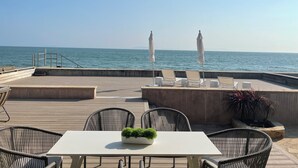 Terraza de madera con mobiliario exterior, una mesa con planta en maceta y dos sombrillas con vistas al mar.