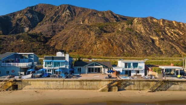 Un hotel costero con varias construcciones, una playa en primer plano y una montaña al fondo.