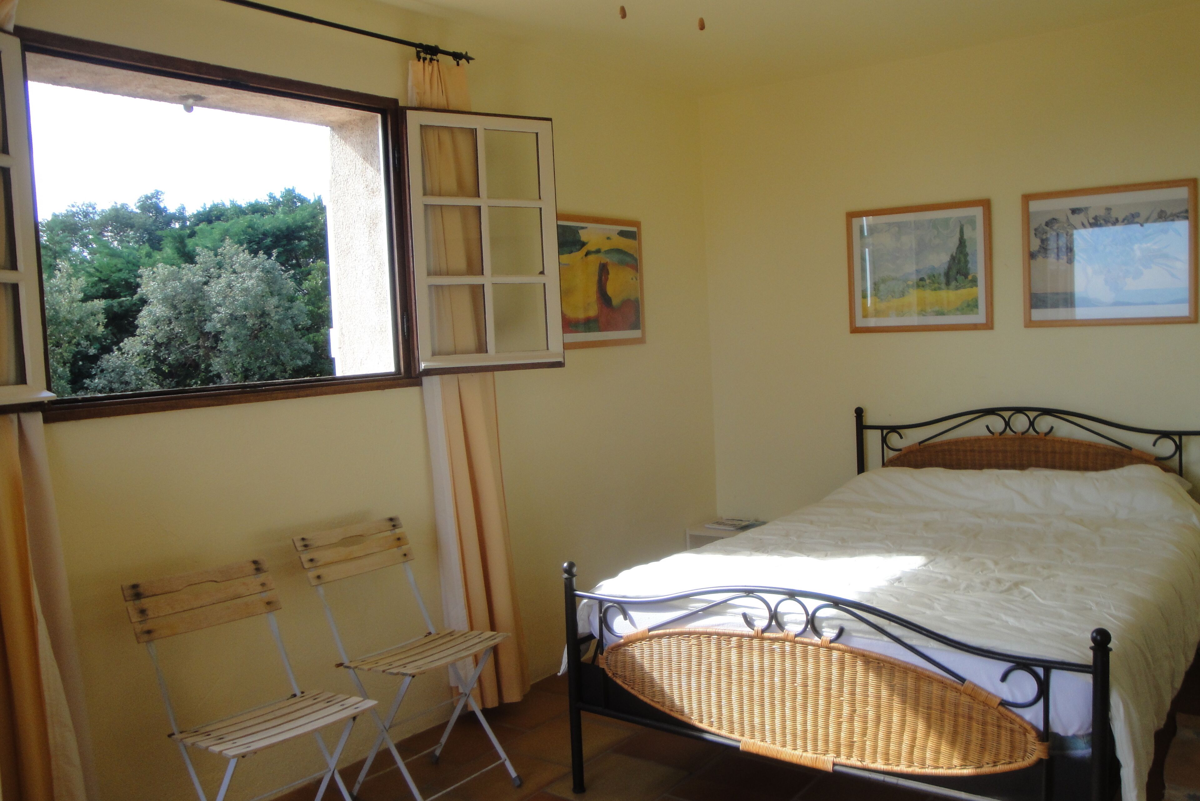 Una camera da letto con un letto, una finestra con tende, due poltrone in vimini e due quadri appesi al muro.
