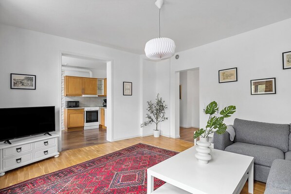 Una sala de estar moderna con un televisor de pantalla plana, un sofá gris, una mesa de centro blanca y una alfombra roja con estampado. Se ve una cocina al fondo con armarios de madera y un microondas.
