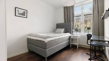 Un dormitorio pequeño y ordenado con cama, escritorio y silla. Hay una ventana con cortinas, un radiador y un cuadro en la pared.