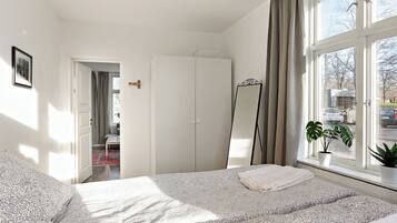A bedroom with a bed, a wardrobe, a mirror, a window with curtains, and a plant.