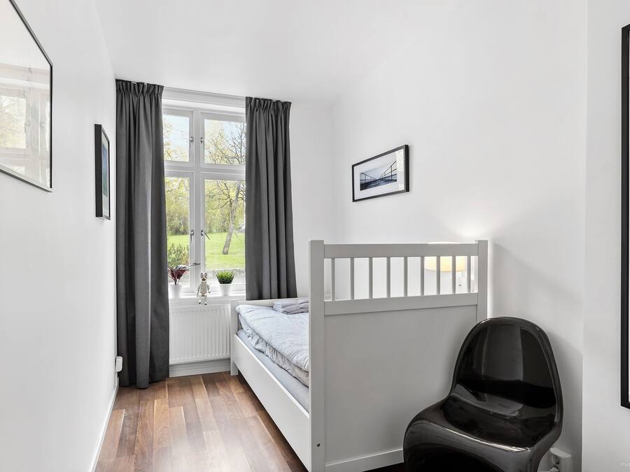 A bedroom with a white bed, a window with curtains, a radiator, and a chair.