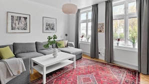 A modern living room with a grey sectional sofa, a white coffee table, and a red patterned rug. There are two framed black and white photographs on the wall, and a window with grey curtains. - HernöRum - Franzéngatan (Harnosand)