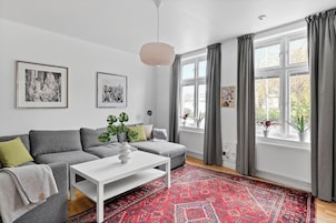 Una sala de estar moderna con un sofá seccional gris, una mesa de centro blanca y un tapete rojo estampado. Hay dos fotografías blanco y negro enmarcadas en la pared y una ventana con cortinas grises.