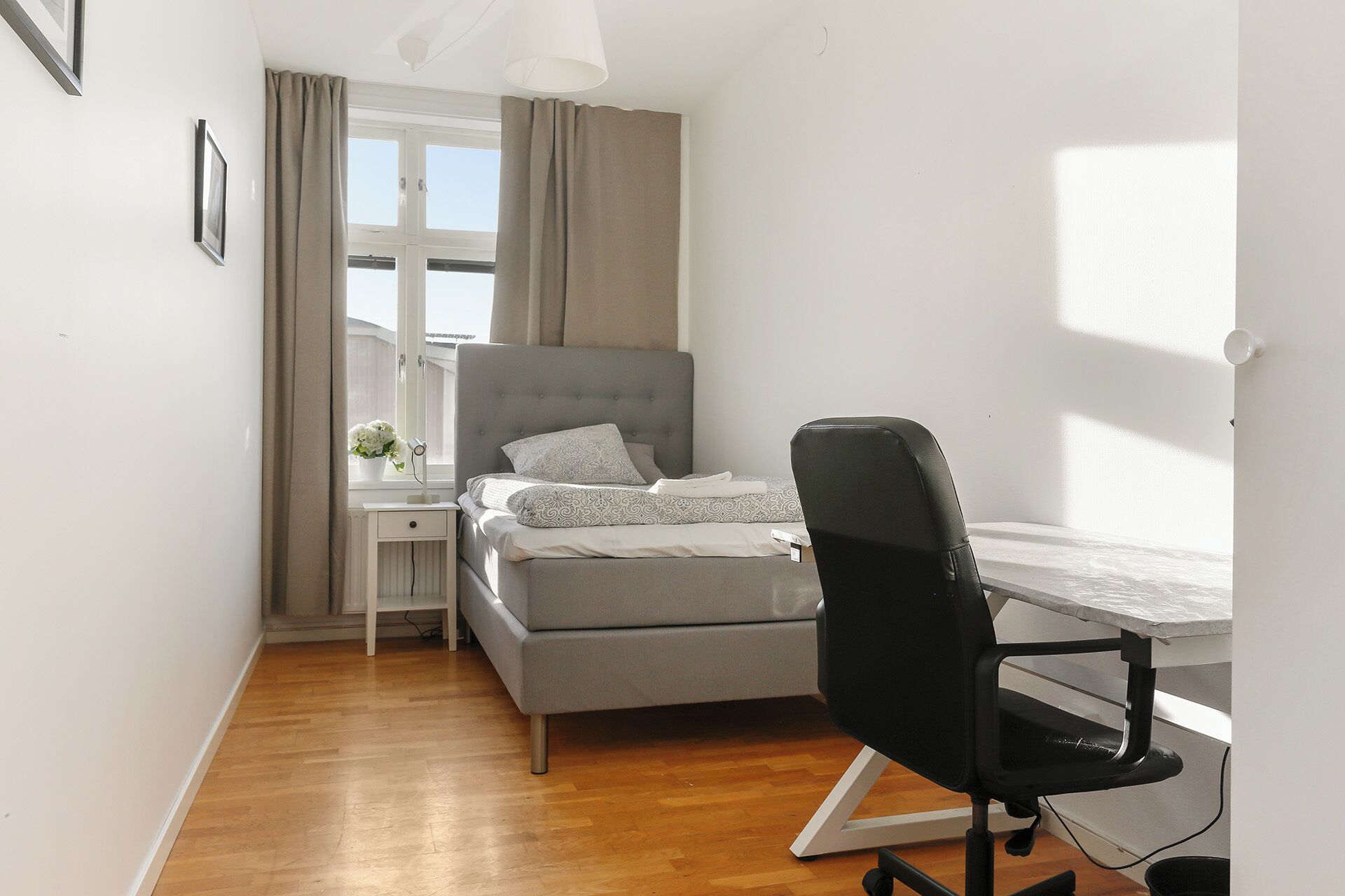 Una camera da letto con un letto, una scrivania, una sedia, una finestra con tende e un comodino con una lampada.