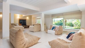A spacious living room with beige sofas, a flat-screen TV, and large windows offering a view of greenery. - Bz73 Geribá! Casa c/ piscina privativa 5 Suítes! (Búzios)